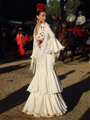 Modelo en la feria de Sevilla con vestido de flamenca blanco roto de 2 volantes grandes y matoncillo blanco roto con flor roja y flor e la cabeza roja