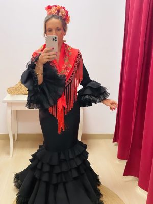 chica vestida con traje de flamenca negro con volantes voluminosos en la falda y mantoncillo coral bordado, y flor en la cabeza